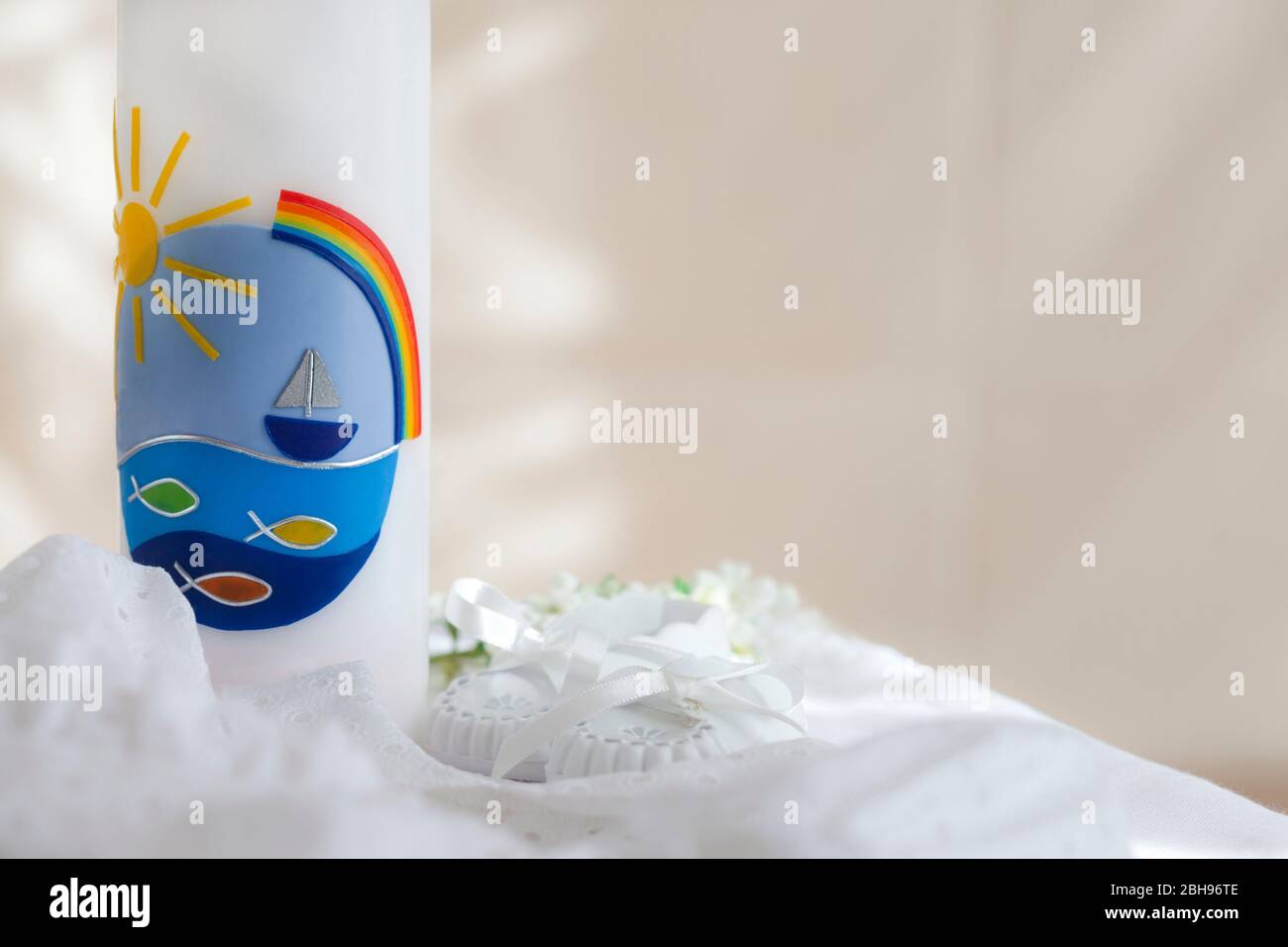 Christening candle with fish, ship and rainbow motif, christening gown and white baby shoes Stock Photo