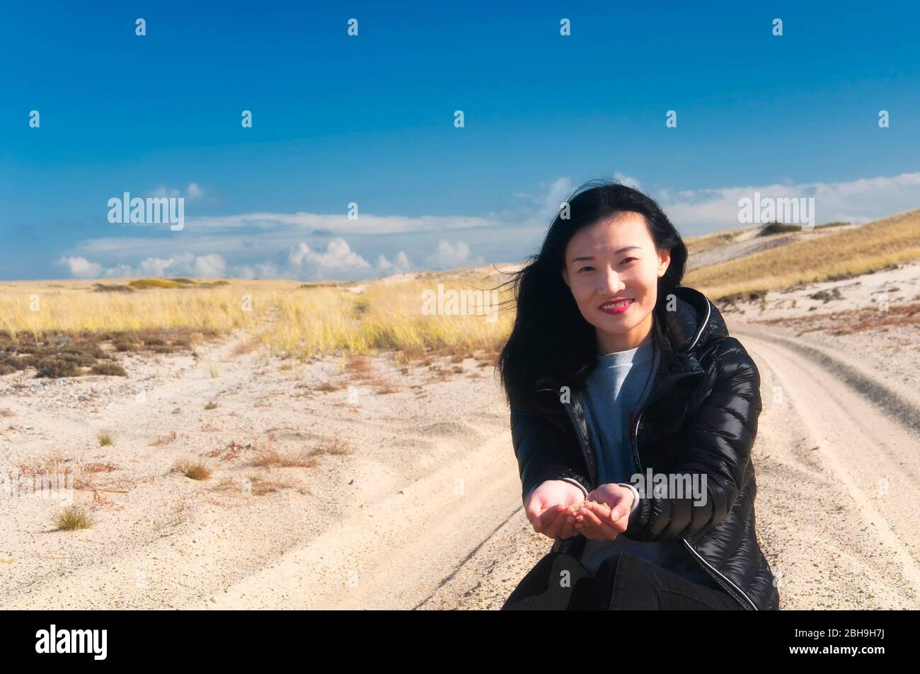 A chinese woman sitting on the sand between tire tracks within the national seashore scenic area in cape cod massachusetts on a sunny blue sky day. Stock Photo