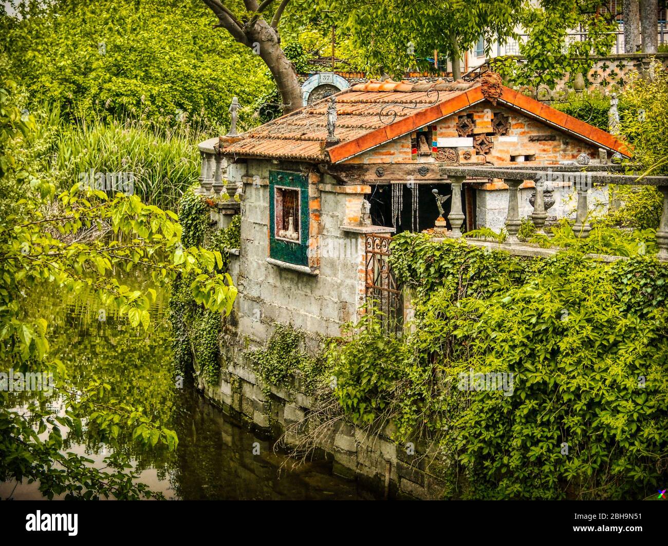 Tiny house on the bank of a stream surrounded by green and luxuriant nature. Statuettes and pottery decorate the exterior. Mantua, Lombardy, Italy. Stock Photo