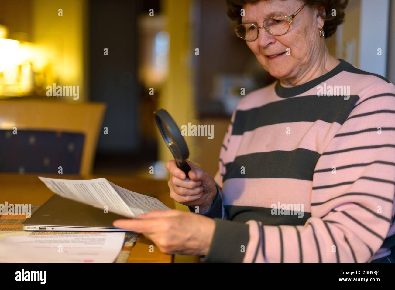 Happy senior woman smiling while reading paper with magnifying glass in the dining room Stock Photo