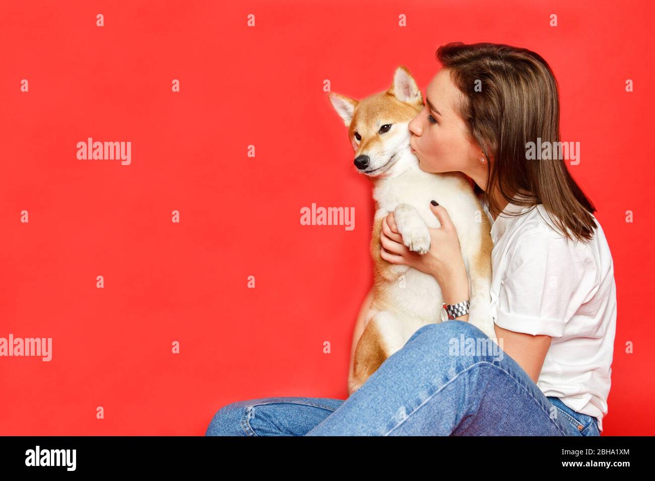 Cute brunette woman in white t shirt and jeans embracing and kissing Shiba Inu dog on plane red background, copy space. Love to the animals, pets conc Stock Photo