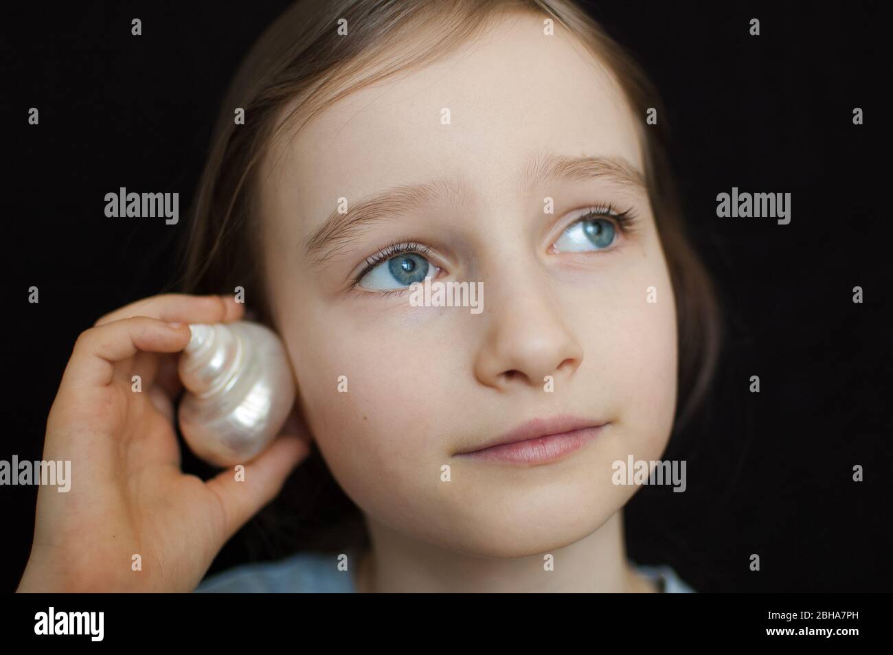 Cute smiling blonde girl holding seashell next to her ear and listening to the ocean on black background in studio Stock Photo