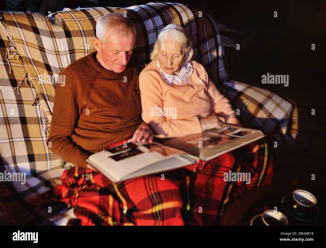 elderly married couple-a man and a woman in a cozy house sitting on the sofa and wrapped in a warm plaid plaid hug each other and smile while viewing Stock Photo