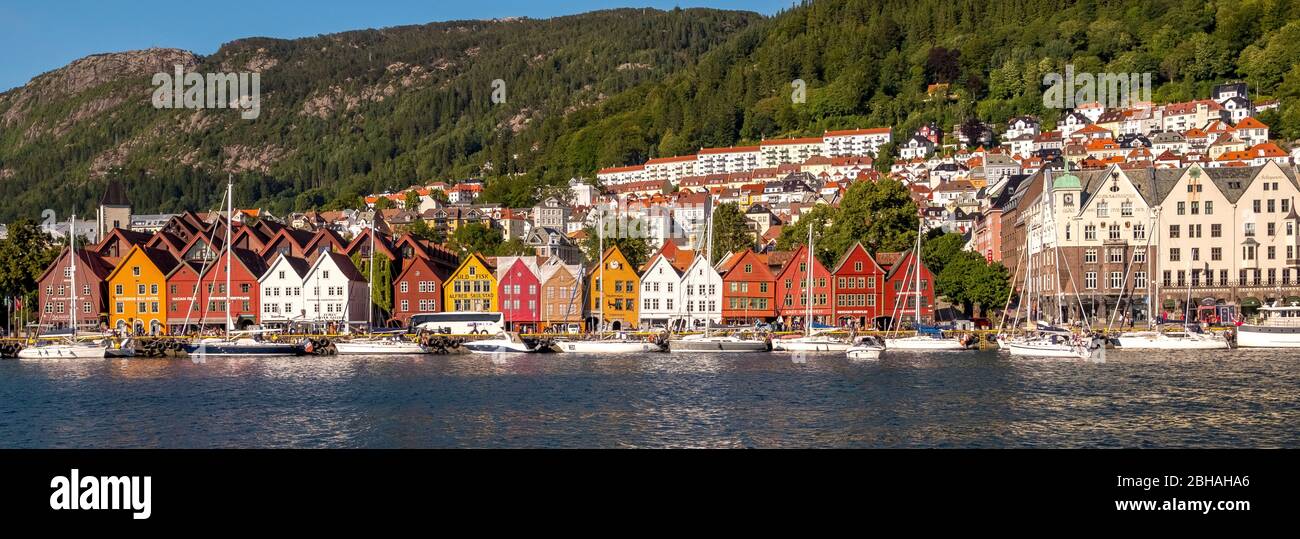 Nordsee, behind a colorful wooden front with attached sailboats in the harbor of Bryggen, behind it more houses of the place above hotel complexes and forest-covered mountains with a piece of bright blue sky, mountains, Hordaland, Norway, Scandinavia, Europe Stock Photo