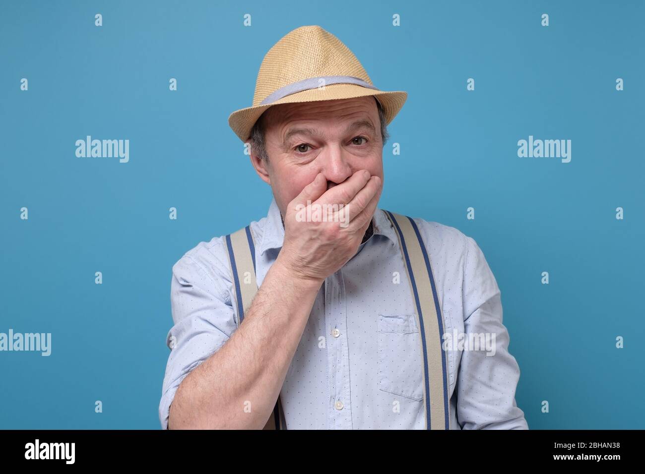 Stunned elderly man in summer hat covers mouth with hands, being surprised Stock Photo
