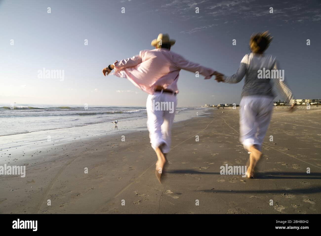 Couple from behind in maritime clothes, holding hands, runs in the surf area of the beach, cheerfulness Stock Photo
