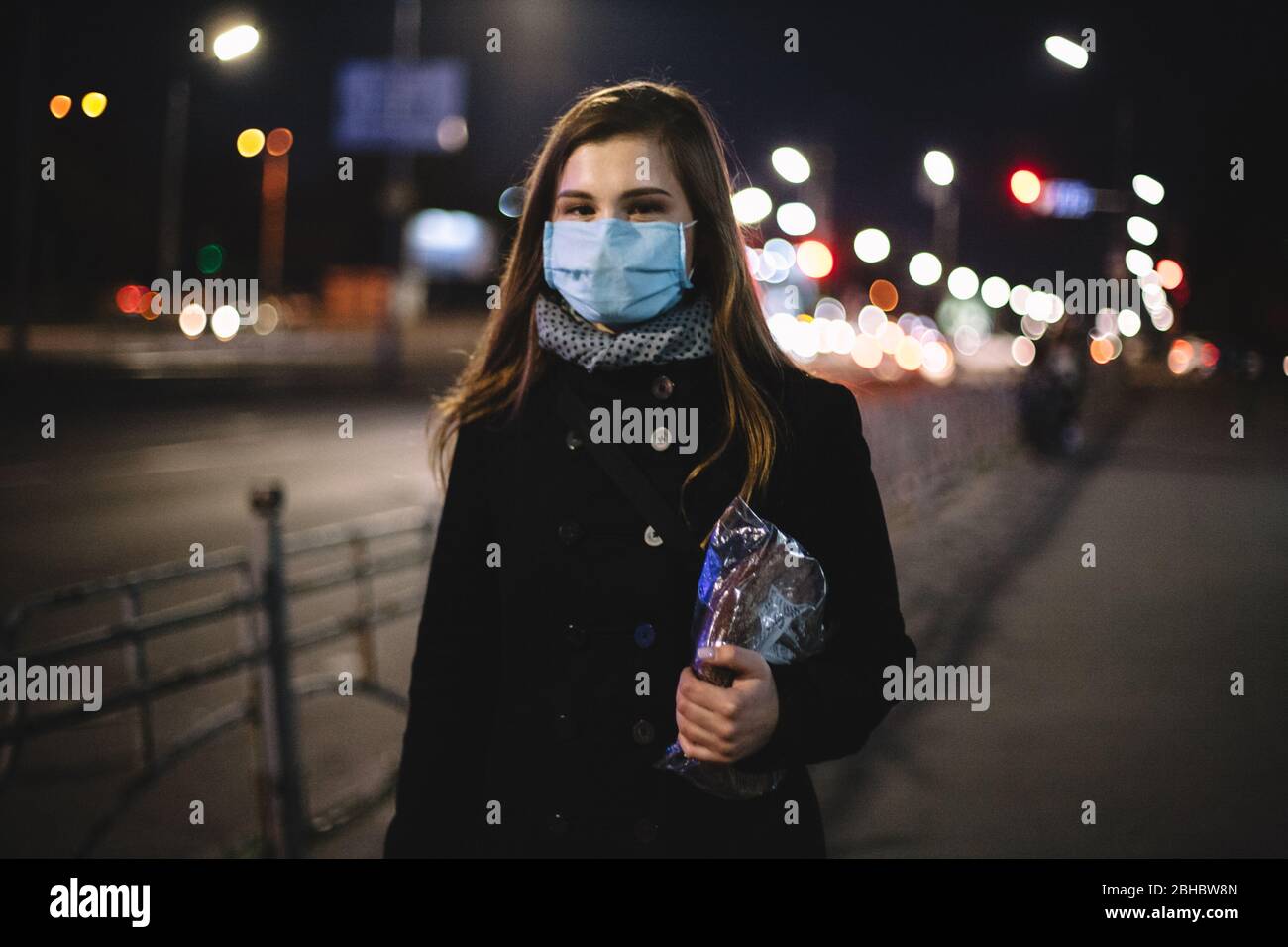Portrait of young happy woman wearing face medical mask carrying bread while walking on city street at night Stock Photo
