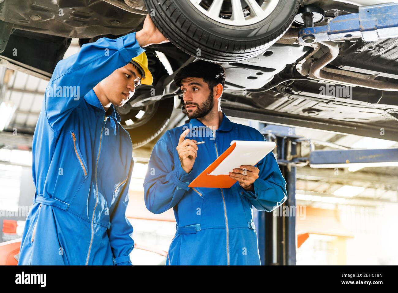 Mechanic in blue work wear uniform inspects the car bottom with his assistant. Automobile repairing service, Professional occupation teamwork. Stock Photo