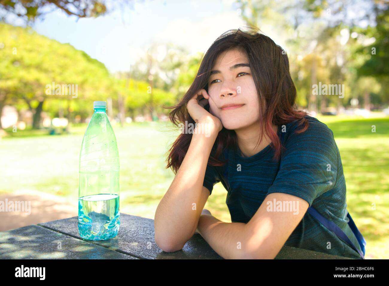 Biracial Asian teen girl sitting outdoors at picnic table in grassy park , looking in the distance, thinking on hot summer day Stock Photo