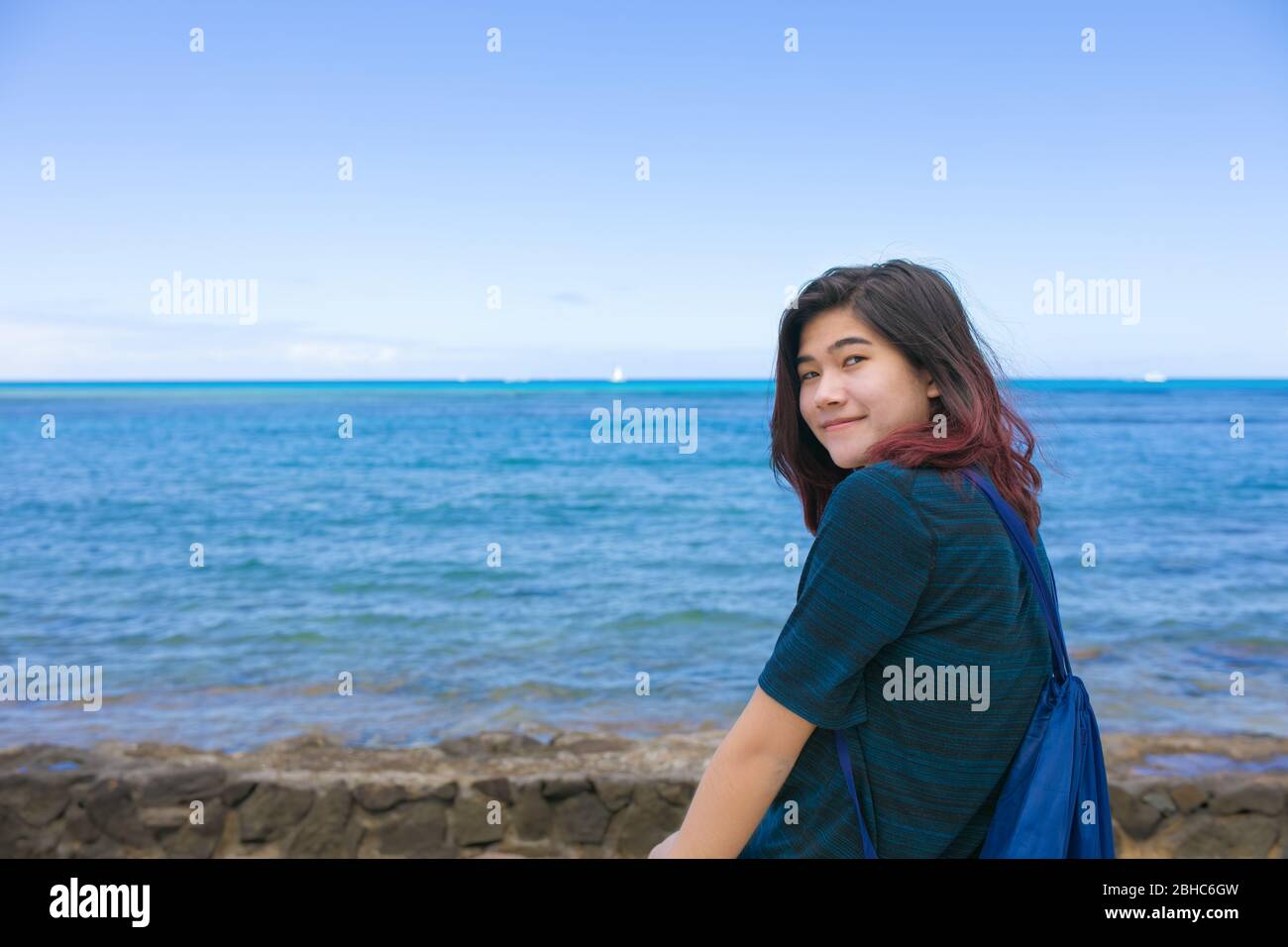Smiling biracial Asian teen girl sitting alone along Hawaiian ocean resting while looking at water Stock Photo