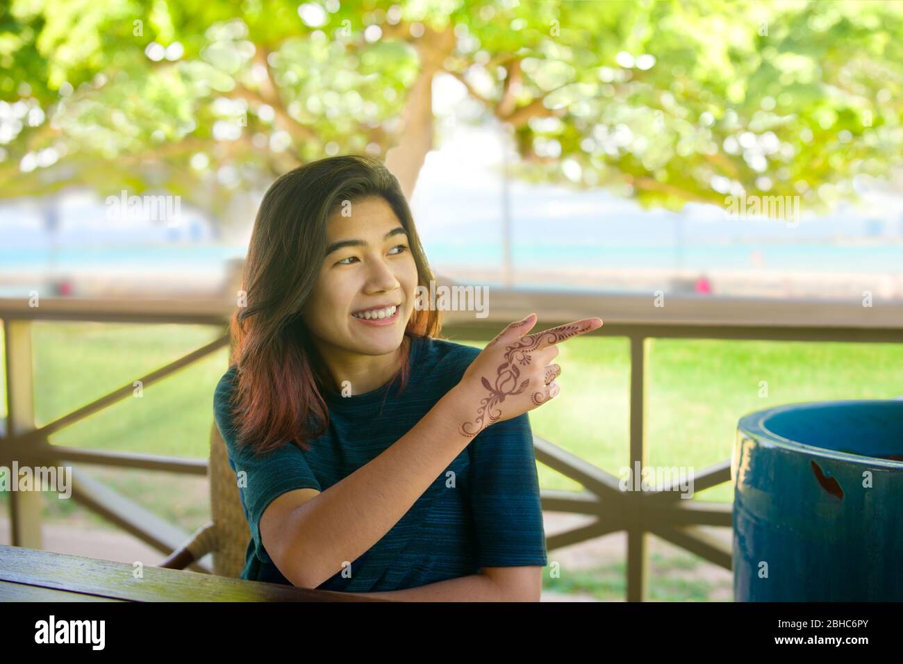 Smiling biracial Asian teen girl smiling and pointing with henna applied hand to something to her left while sitting in an outdoor restaurant with sun Stock Photo