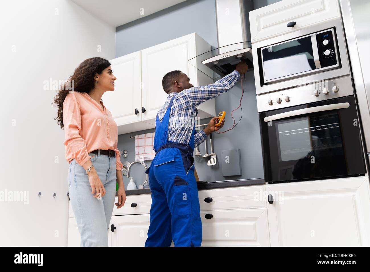 Male Repairman Checking Extractor With Digital Multimeter While Woman Standing In Kitchen Stock Photo