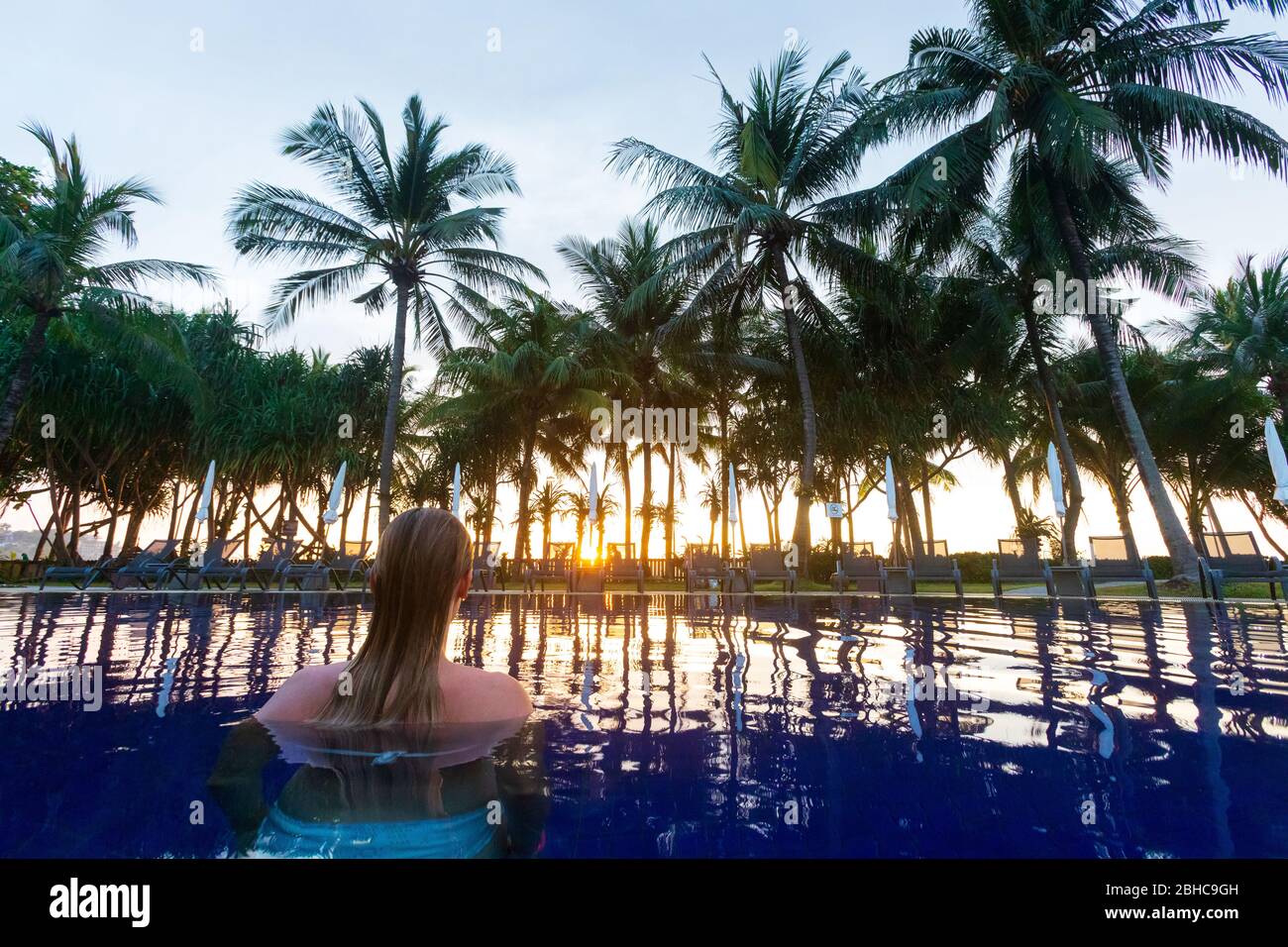 Vacation in a tropical exotic resort by the pool. A woman back inside a swimming pool with sunset lights. Stock Photo