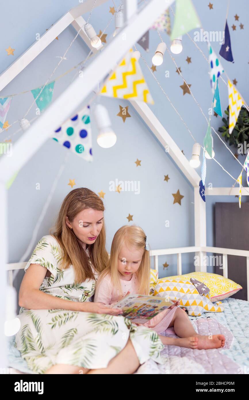 Closeup of pretty 3-year-old girl and her young mother reading a book in bed Stock Photo