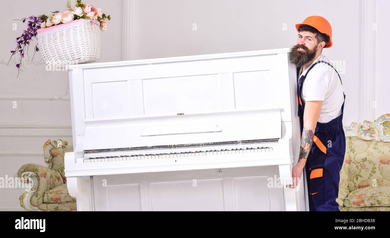 moves piano instrument. Delivery service concept. Courier delivers furniture in case of move out, relocation. Man with beard, worker in overalls and lifts up piano, white background Stock Photo -