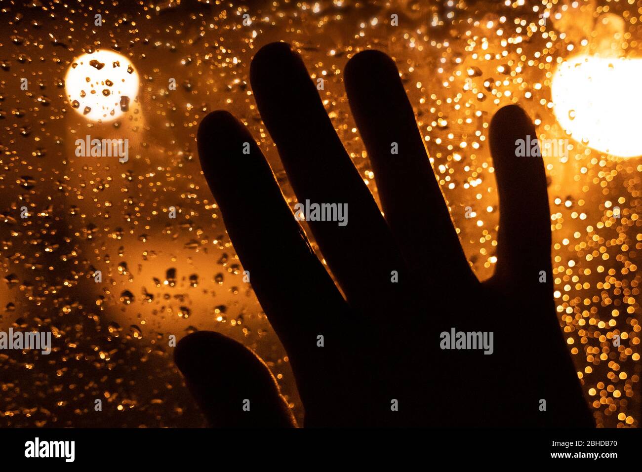Silhouette of a hand leaning against the window at night with drops of water. Sad image of melancholy looking out from the sleeping room. Stock Photo