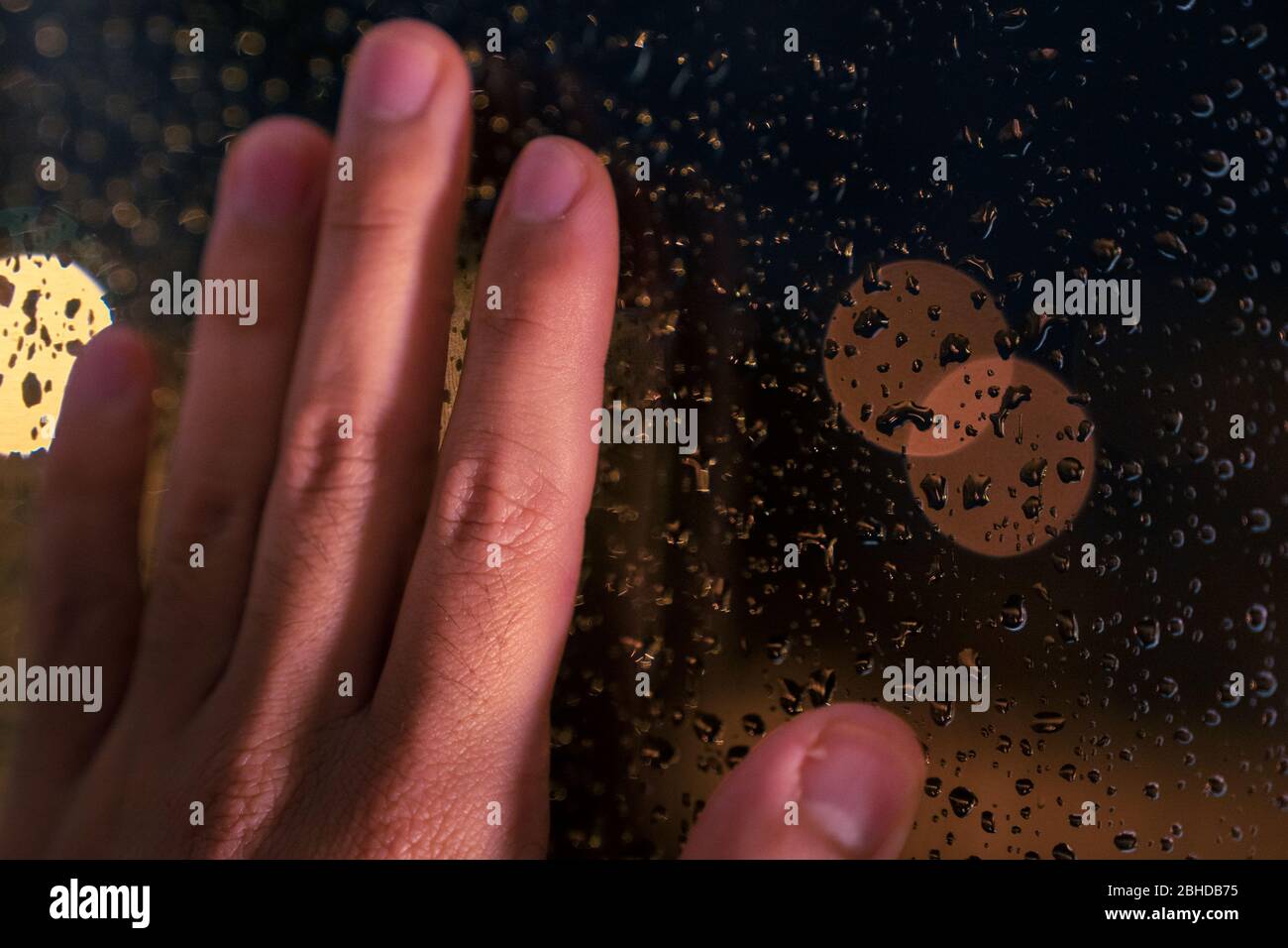 Hand resting on the window at night with the warm light of the street lamps and with drops of water from a rainy day. Sad image of melancholy looking Stock Photo