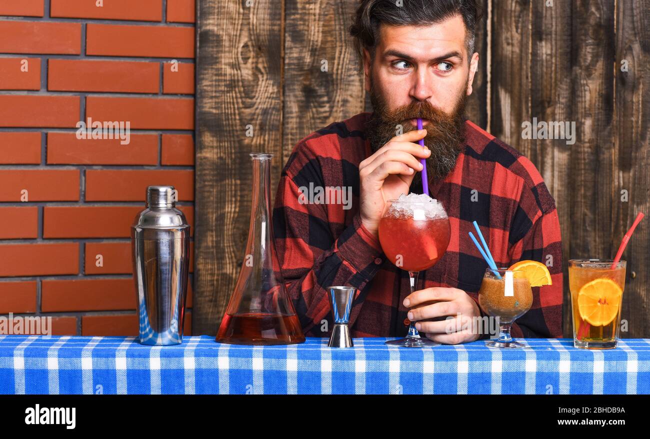Barman with beard and surprised face drinks out of glass with drinking straw cocktail. Man drinks cocktail on wooden background. Alcohol and cocktails concept. Hipster enjoy drink or cocktail. Stock Photo
