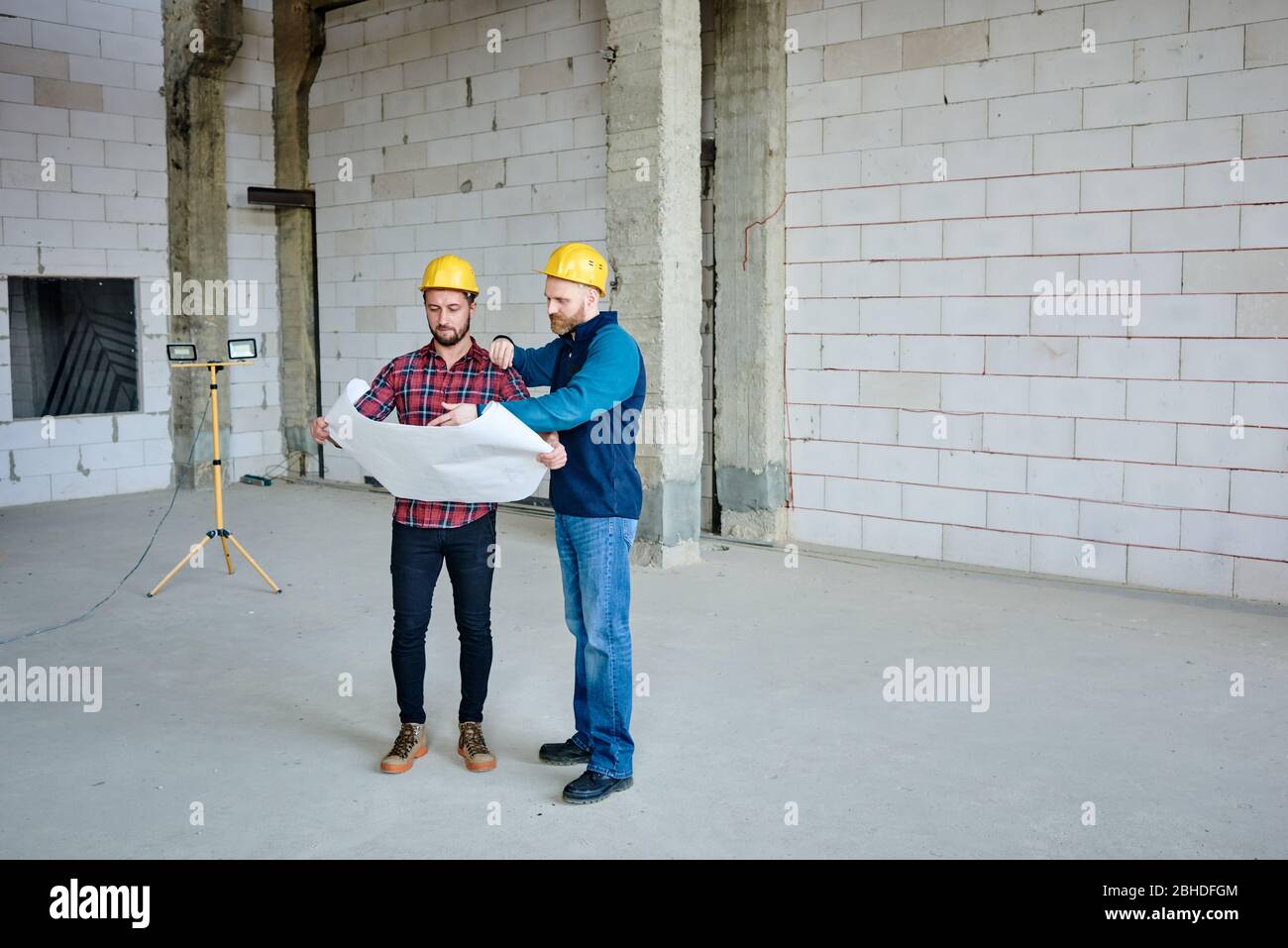 Two young builders in protective helmets discussing sketch in blueprint while pointing out the main structure at start-up meeting Stock Photo
