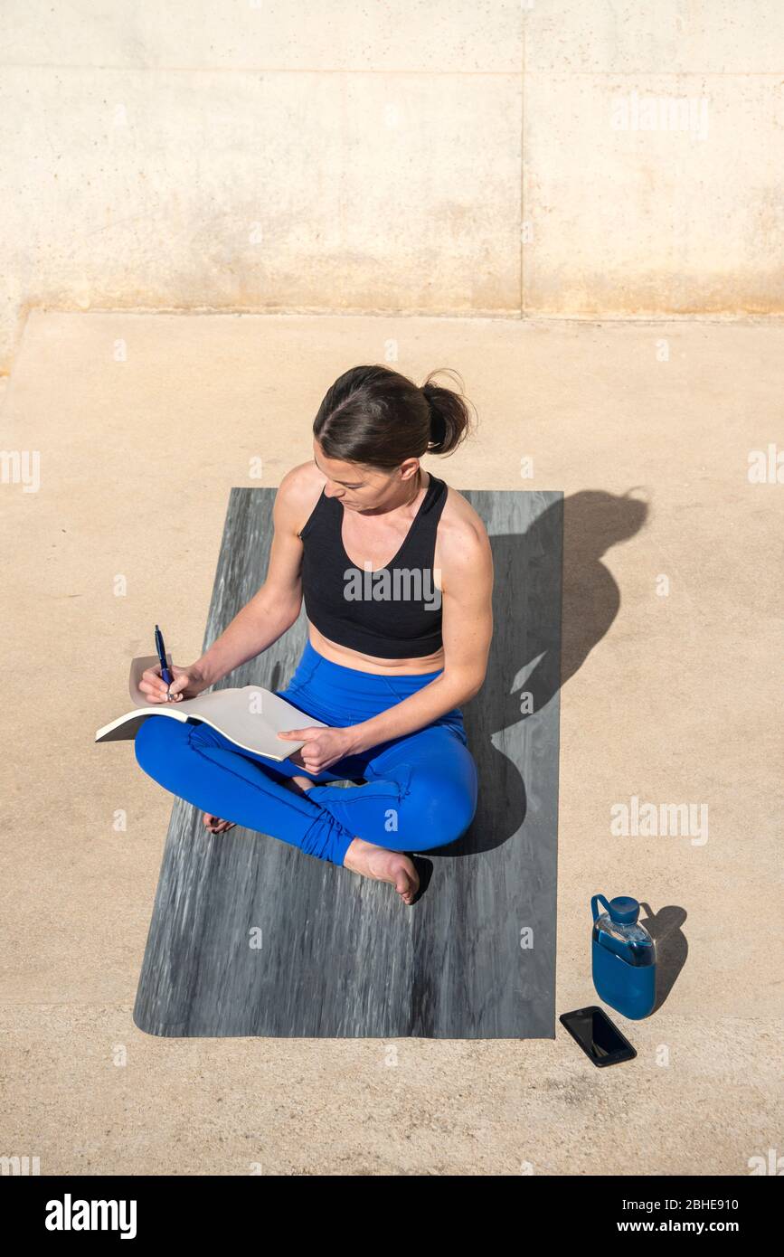 Sporty woman sitting on an exercise mat outside writing in a notebook, journaling. Stock Photo
