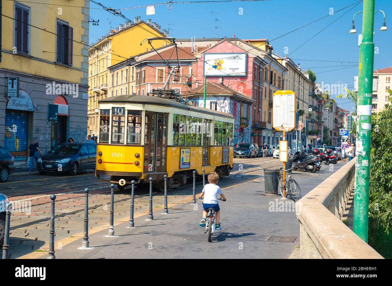 Milan, Italy, September 9, 2018: young boy is riding a bicycle bike near old yellow traditional tram on street with colorful multicolored buildings in beautiful summer day Stock Photo