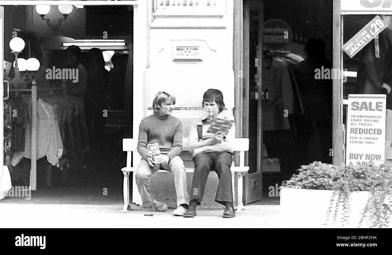 Daily life on the streets in Manchester, England, United Kingdom in 1974. Two young teenage boys sit chatting on a bench outside a clothes shop. Stock Photo