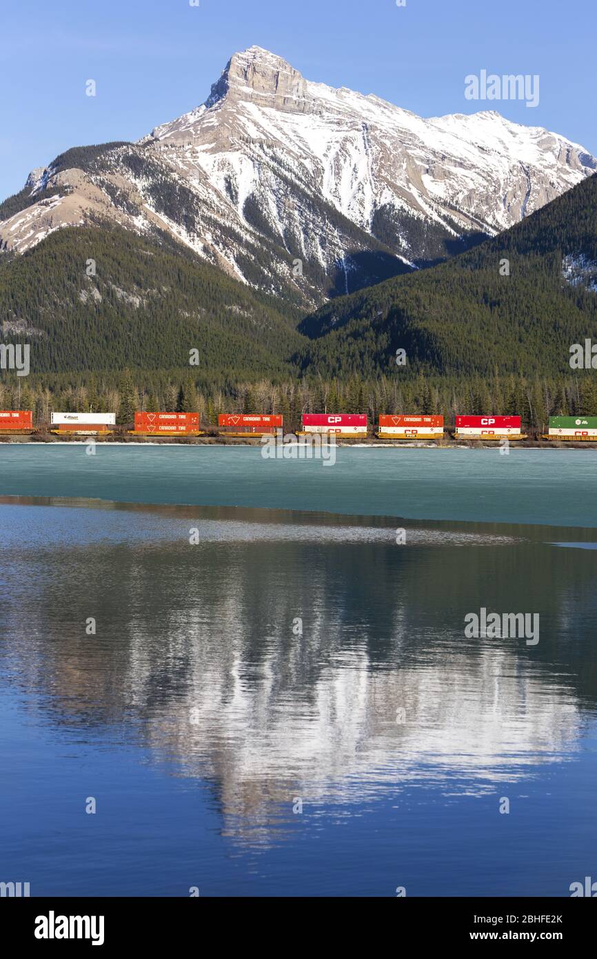 CP Railway Freight Train passing in Alberta Foothills of Canadian Rockies with Snowy Mountain Peaks reflected in calm water of Gap Lake Stock Photo