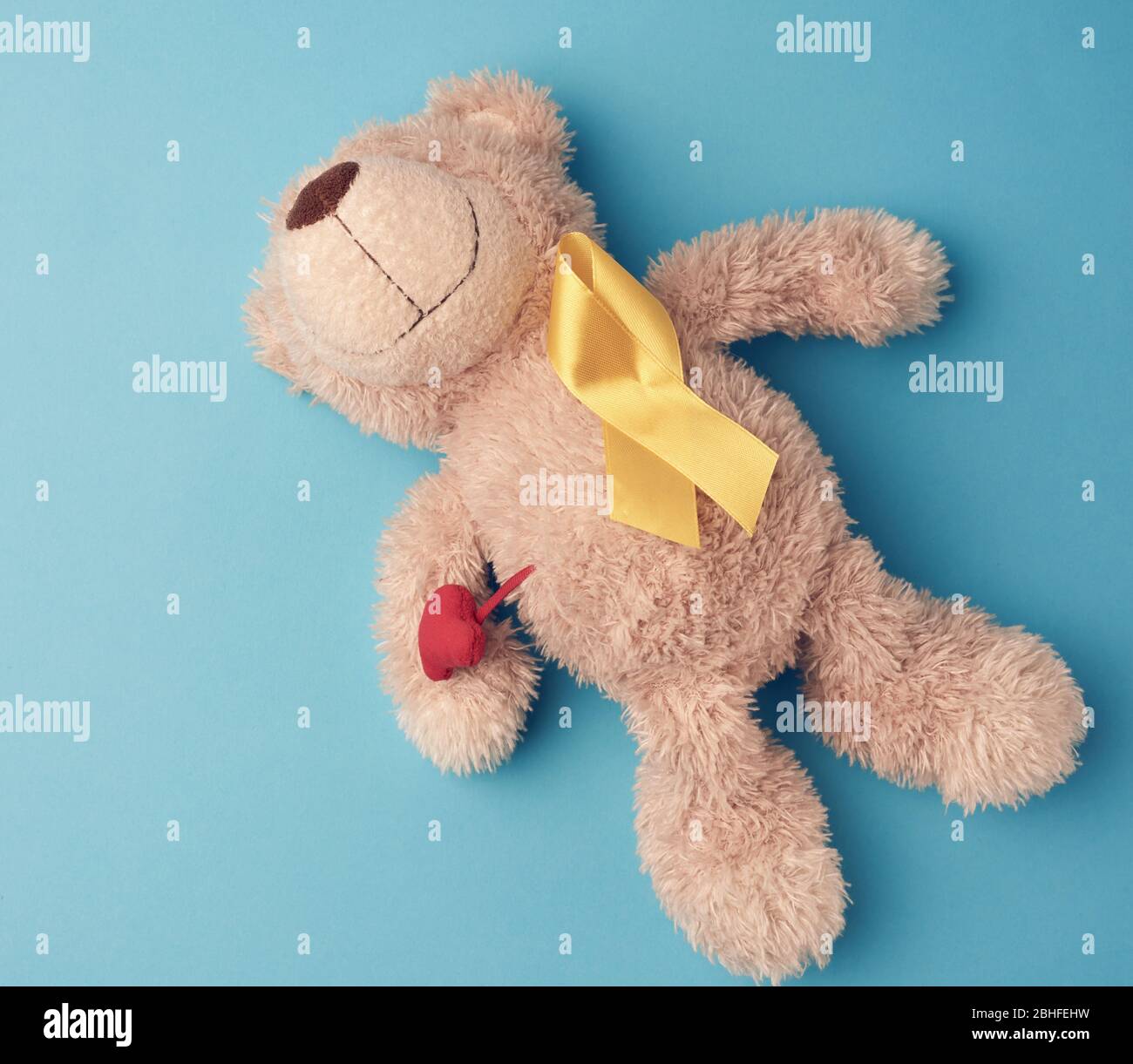 teddy bear holds in his paw a yellow ribbon folded in a loop on a blue background. concept of the fight against childhood cancer. problem of suicides Stock Photo