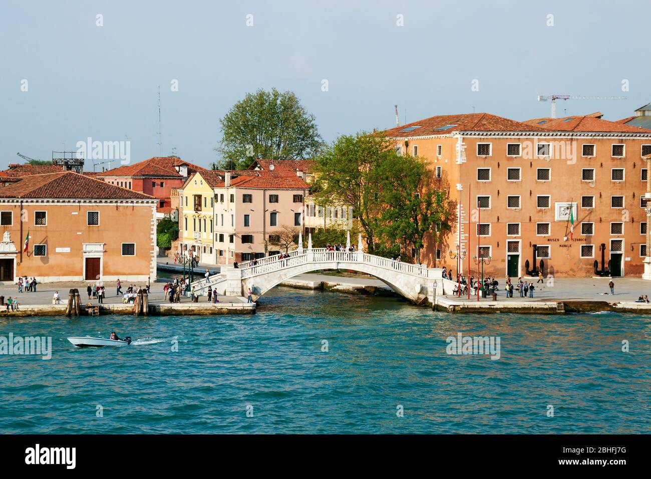 A bridge over the entrance to a small residential canal from the Grand Canal. People walking and riding in a motorboat. Stock Photo