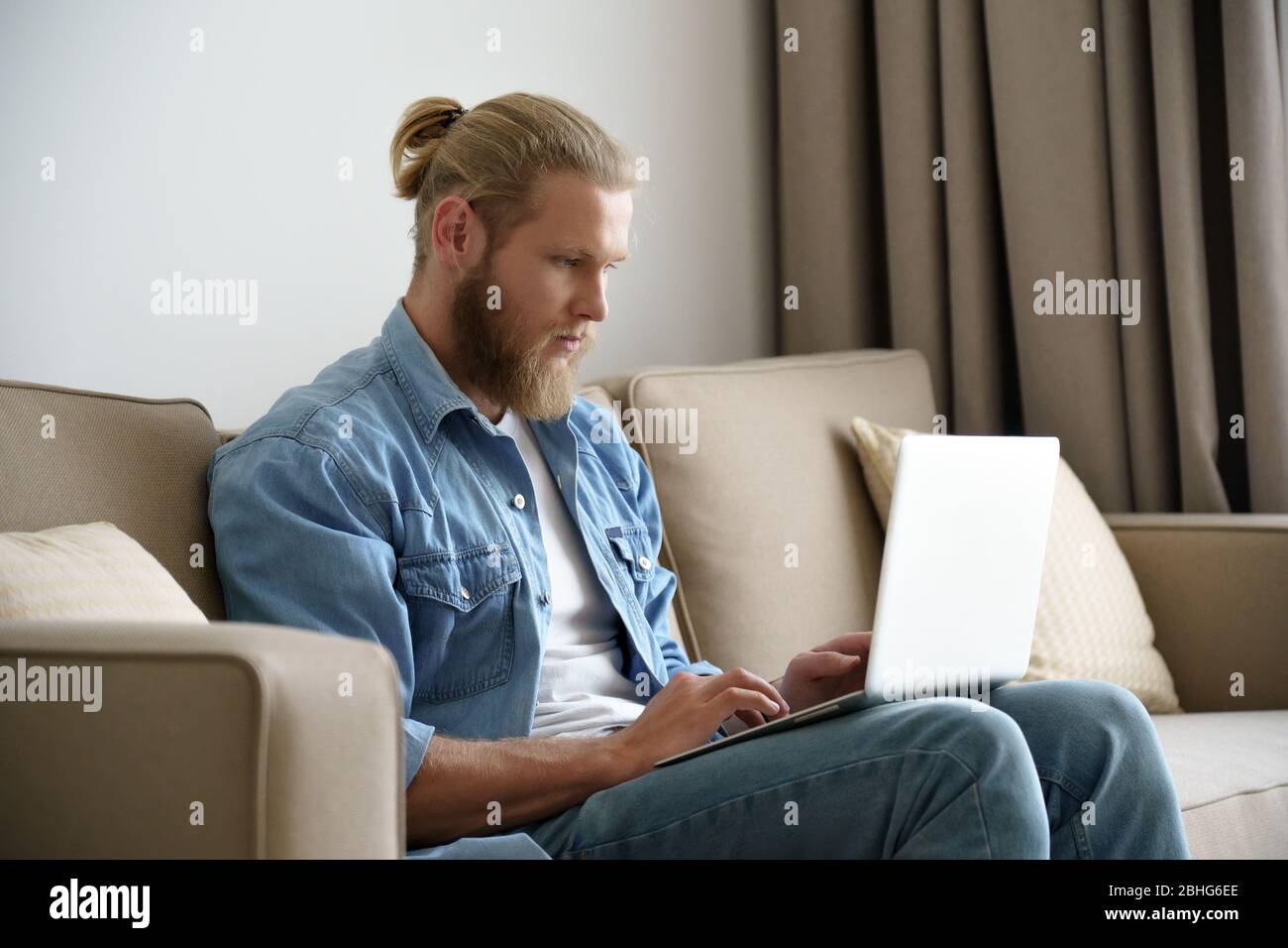 Serious young man working studying from home using laptop sitting on couch. Stock Photo