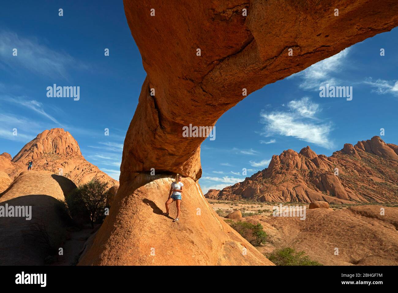 Girl under natural rock arch at Spitzkoppe (left), and Pondok Mountains in distance (right), Namibia, Africa Stock Photo
