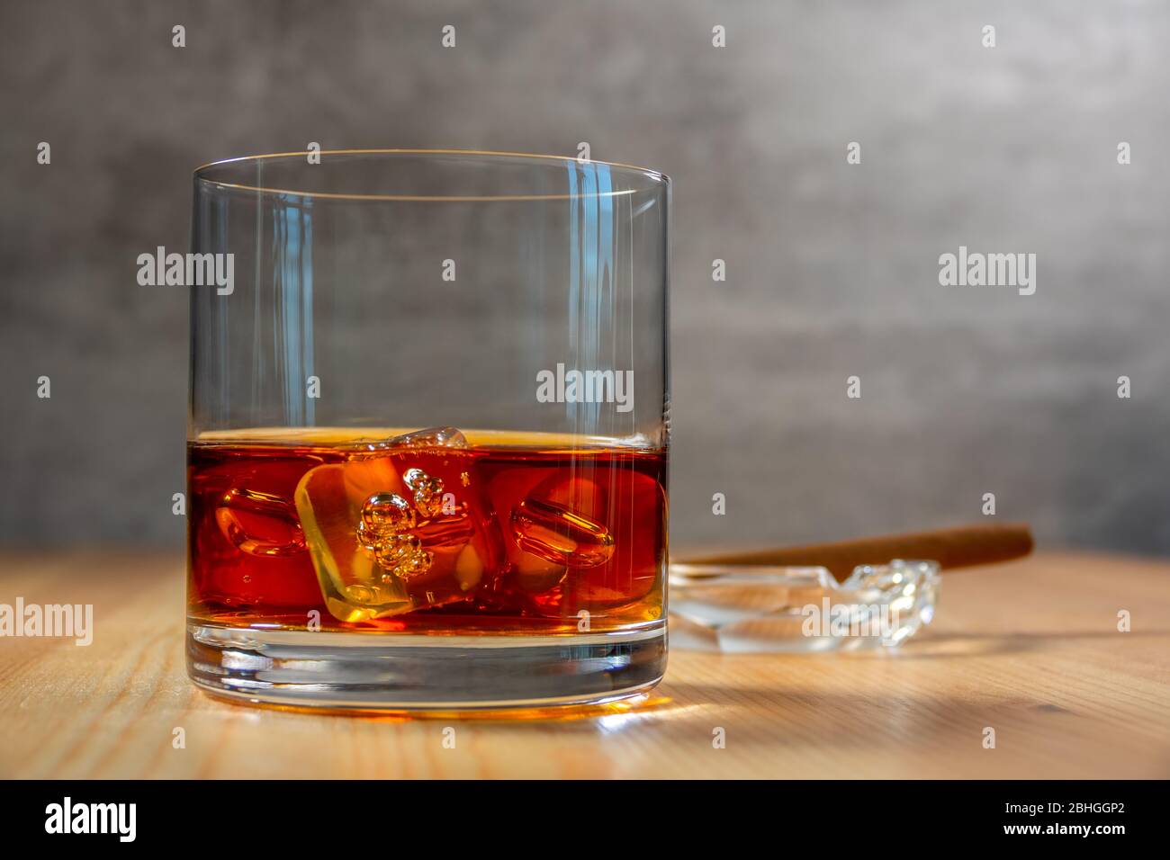 Ashtray with a cigar in the background in defocus. Glass of whiskey with ice cubes on the wooden table Stock Photo