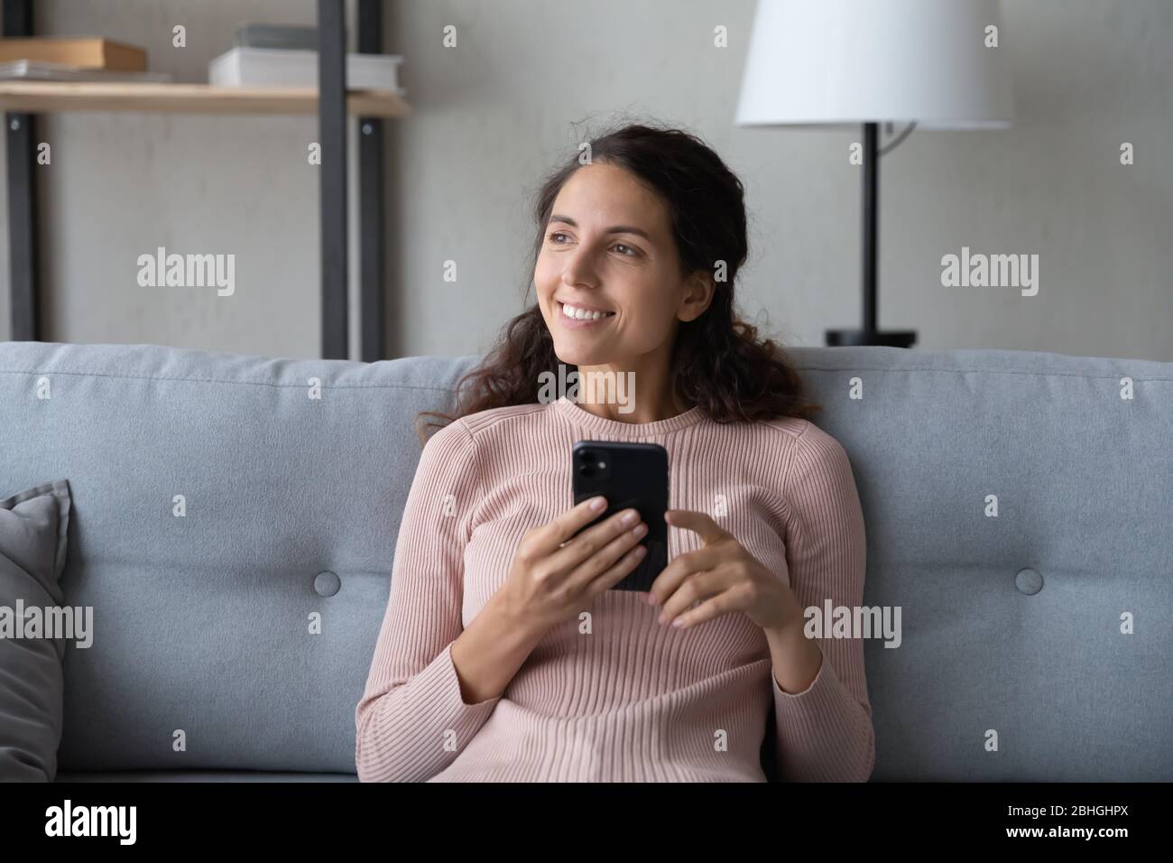 Distracted young smiling woman resting on sofa, holding smartphone. Stock Photo