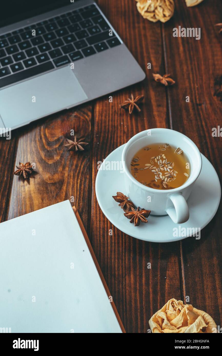White cup of tea with star anise with a notebook on a wooden table with a computer in the background Stock Photo