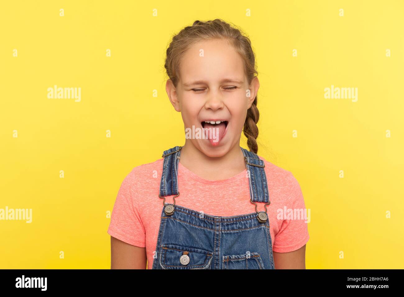 Portrait of naughty little girl in denim overalls sticking out tongue and keeping eyes closed, disobedient kid fooling around with funny derisive expr Stock Photo