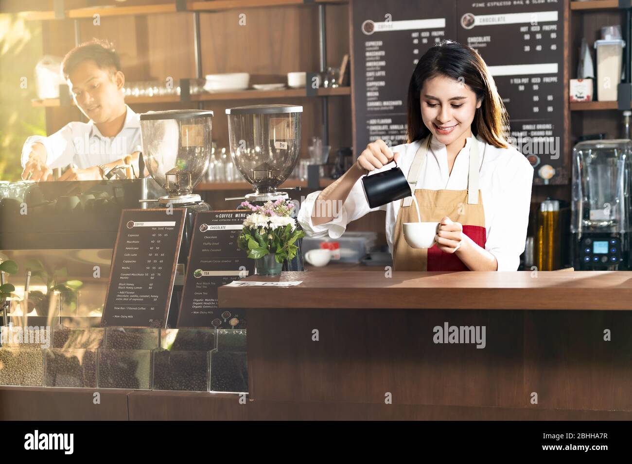 Young adult asian female woman barista pouring fresh milk to prepare latte coffee for customer in cafe bar with her colleague working in background. F Stock Photo