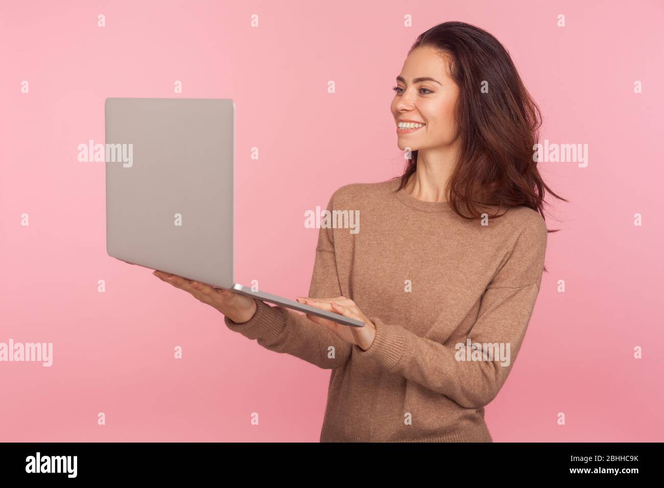 Portrait of young woman in casual sweater standing, holding laptop and smiling while talking on video call, having online conference, freelancer worki Stock Photo