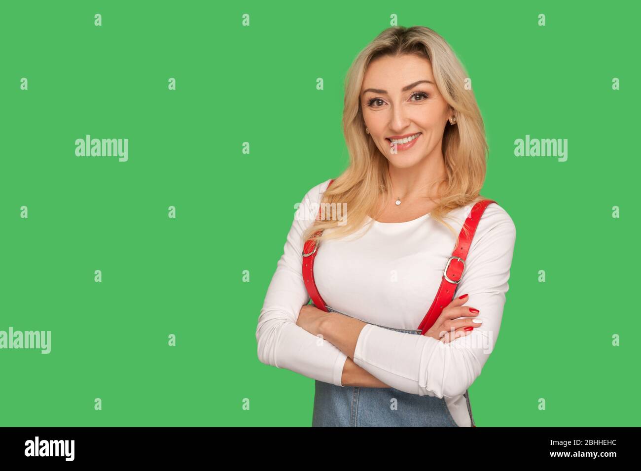 Portrait of positive self assured beautiful adult woman in stylish overalls holding arms crossed, looking at camera with toothy smile and confident ex Stock Photo