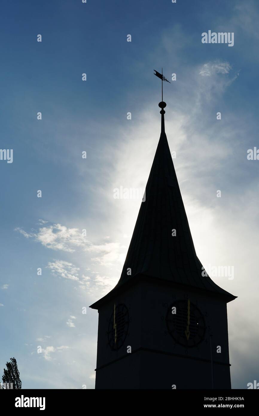 A silhouette of a church tower with a wind pointer on the background of slightly covered sky and surrounded by sunlight Stock Photo