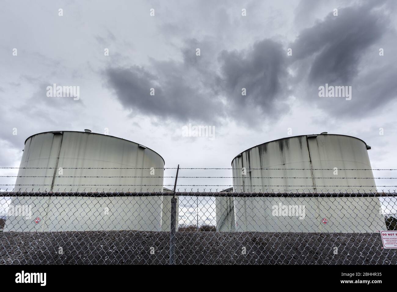Large jet fuel or oil storage tanks behind a chain link fence with warning signs. Stock Photo