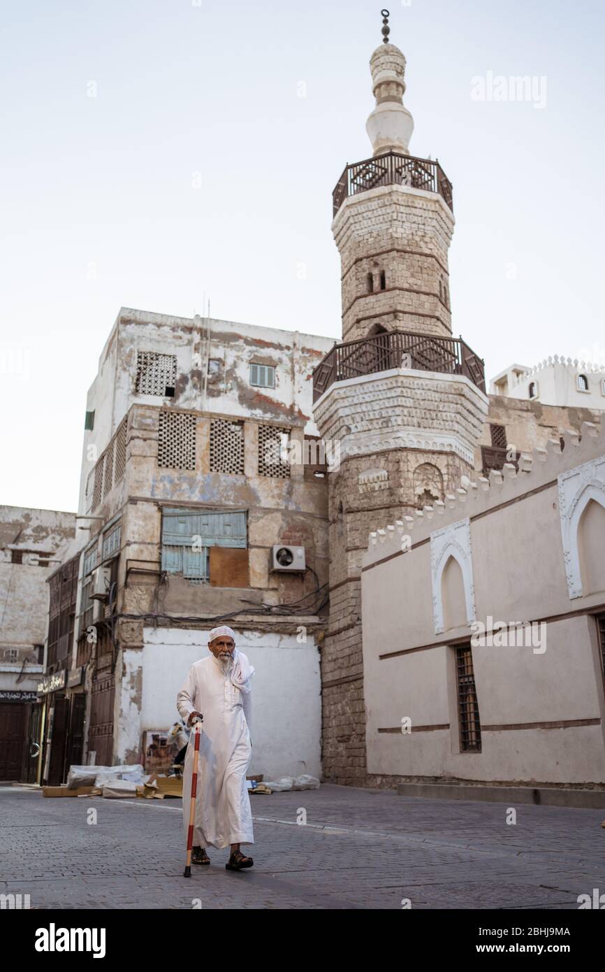 Jeddah / Saudi Arabia - January 16, 2020: Muslim man wearing traditional clothes walking in front of old mosque with minaret in the streets of Al-Balad Stock Photo