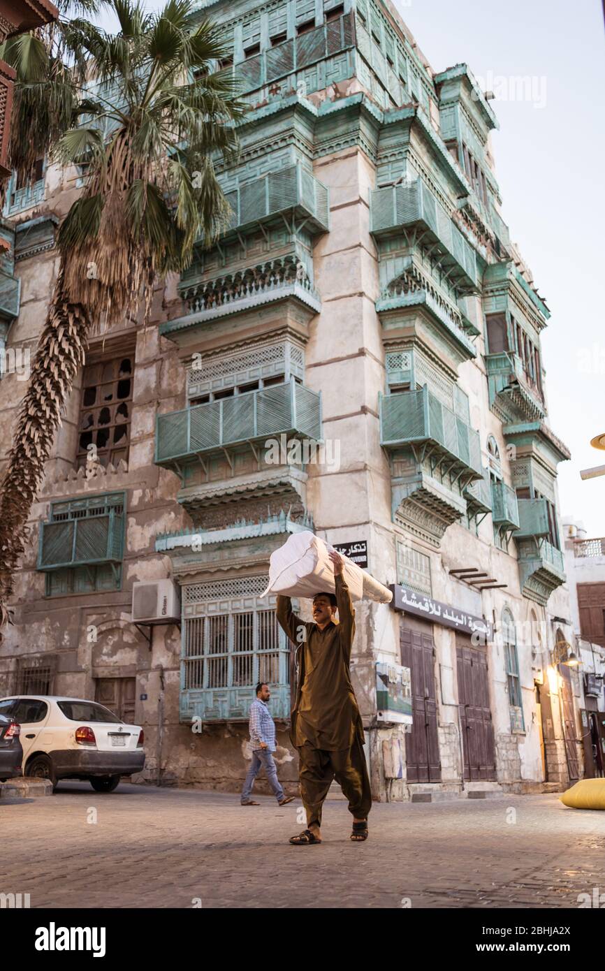 Jeddah / Saudi Arabia - January 16, 2020: Man carrying a package on his head with historic colorful building in Al-Balad in the background Stock Photo