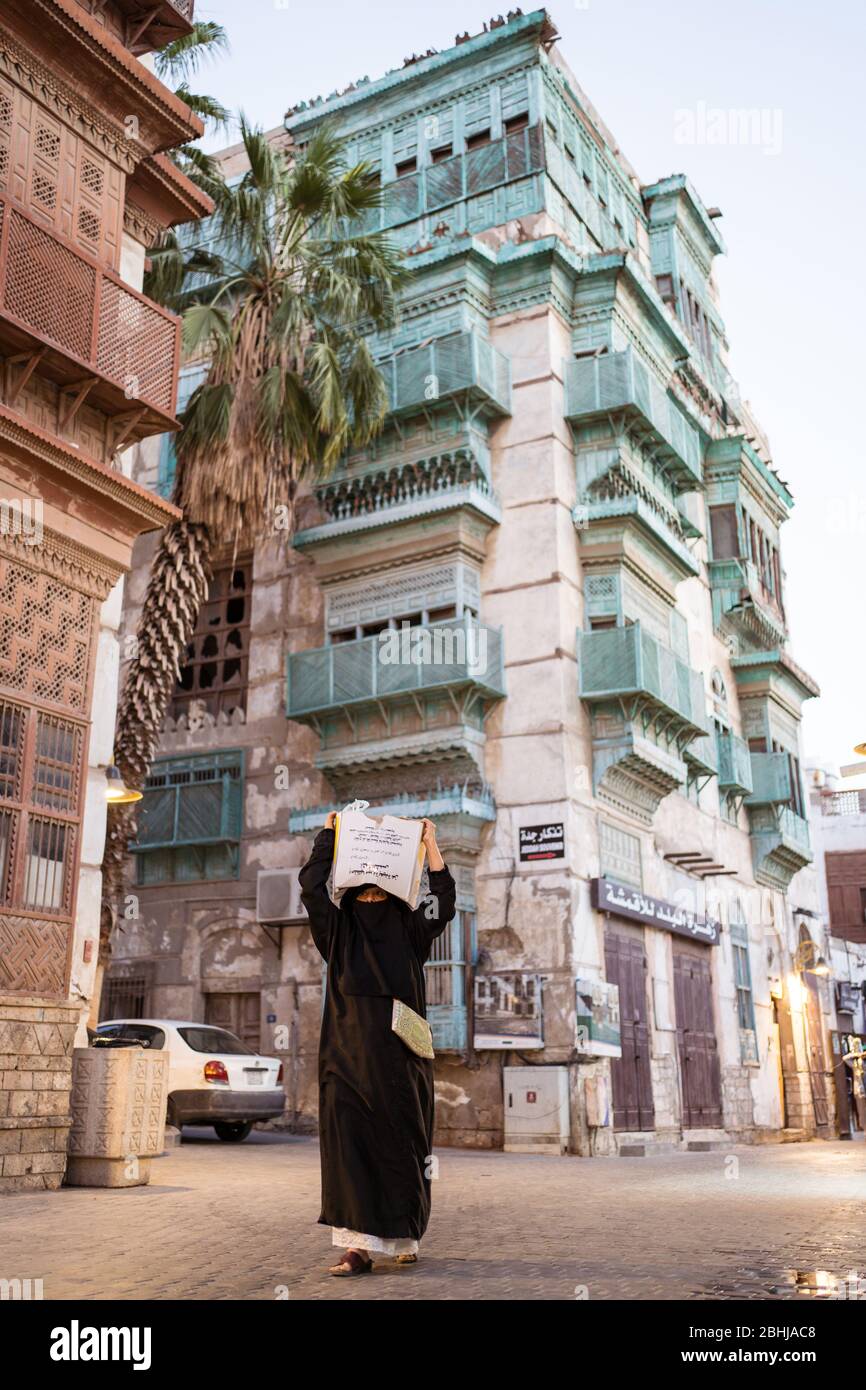 Jeddah / Saudi Arabia - January 16, 2020: Woman in abaya and niqab carrying a package on her head with historic colorful building in the background Stock Photo