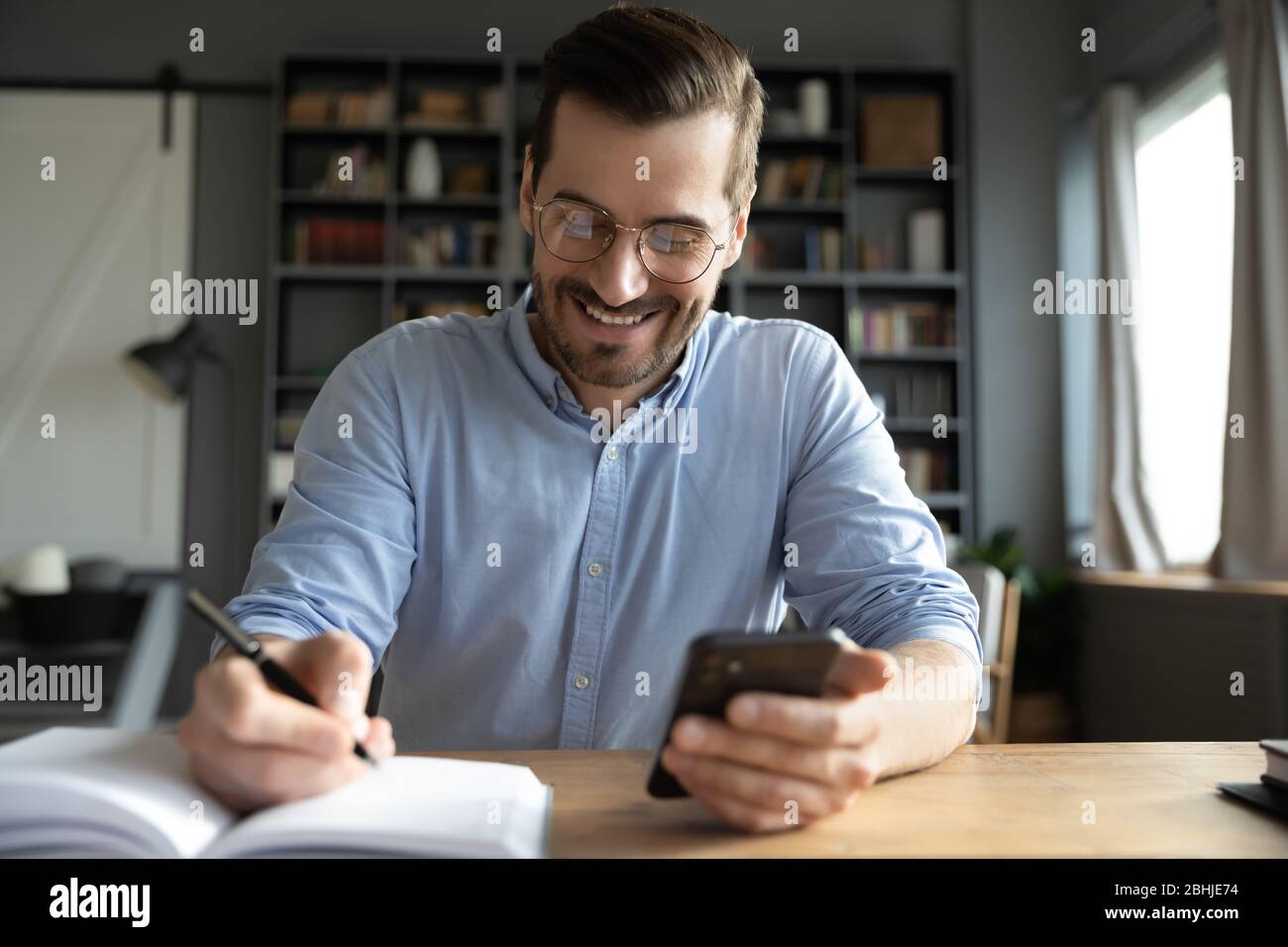 Smiling businessman wearing glasses using phone, writing in notebook Stock Photo