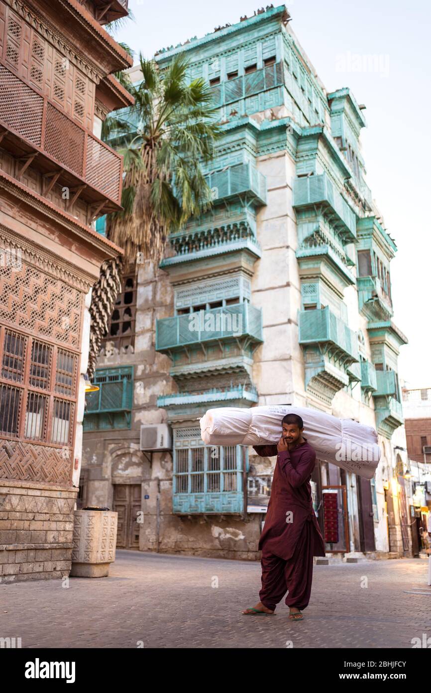 Jeddah / Saudi Arabia - January 16, 2020: Man carrying a package on his head with historic colorful building in Al-Balad in the background Stock Photo