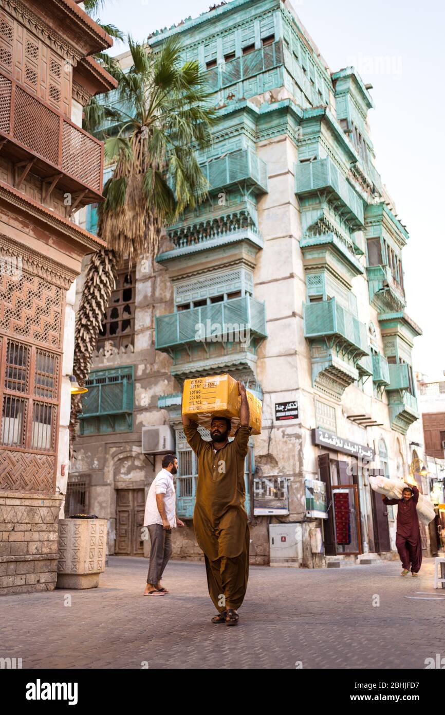 Jeddah / Saudi Arabia - January 16, 2020: Man carrying a package on his head with historic colorful building in Al-Balad in the background Stock Photo