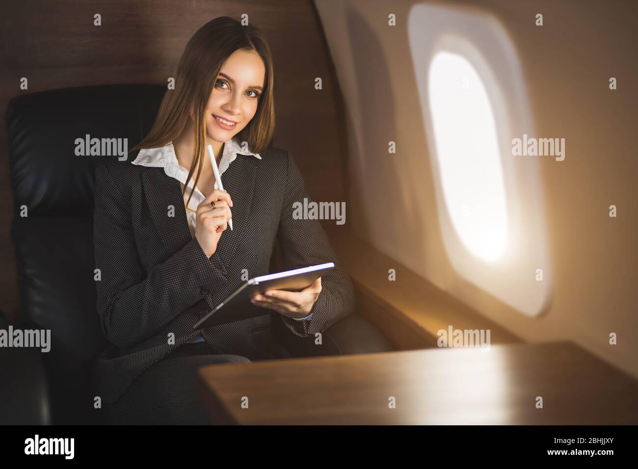 Happy brown haired young caucasian businesswoman holding tablet, sitting on seat in private jet, looking at airplane window and smiling. Stock Photo
