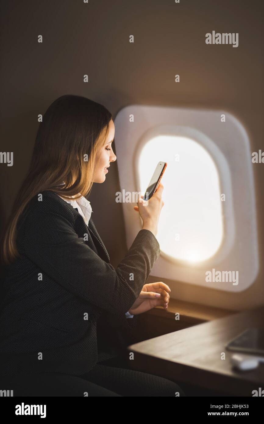 Adorable caucasian girl traveling by private airplane. Charming brown haired young woman sitting by aircraft window and taking pictures of sky on her Stock Photo