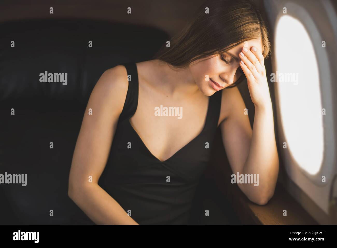 Young woman is afraid to fly. Closeup shot of beautiful tired girl sitting in airplane at window, touching her head, experiencing stress, headache, mi Stock Photo