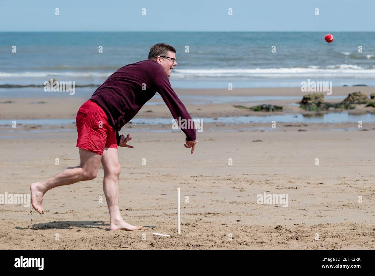 A man on the beach playing cricket, bowling the ball in front of the